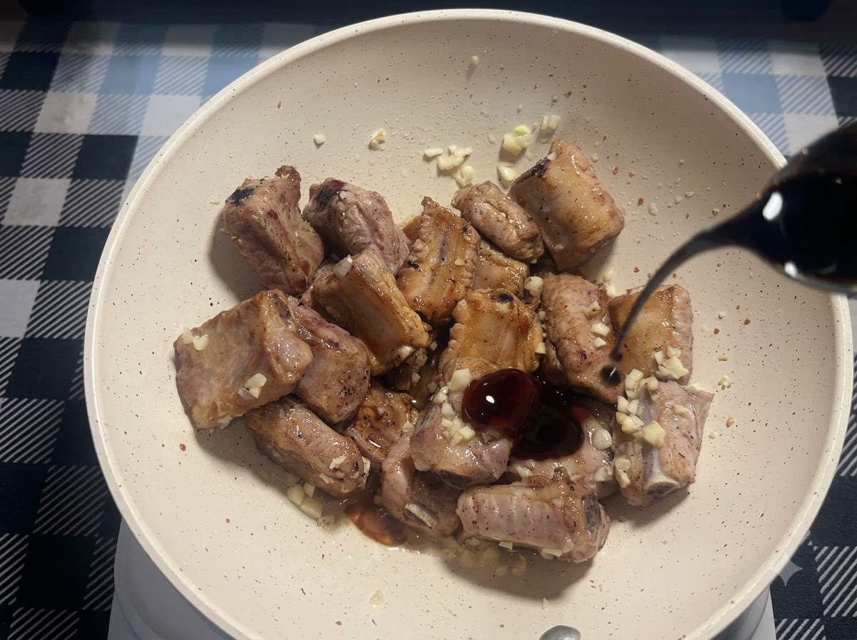 Un petit filet de sauce soja noire versé dans la poêle avec les travers de porc frits et lail haché.