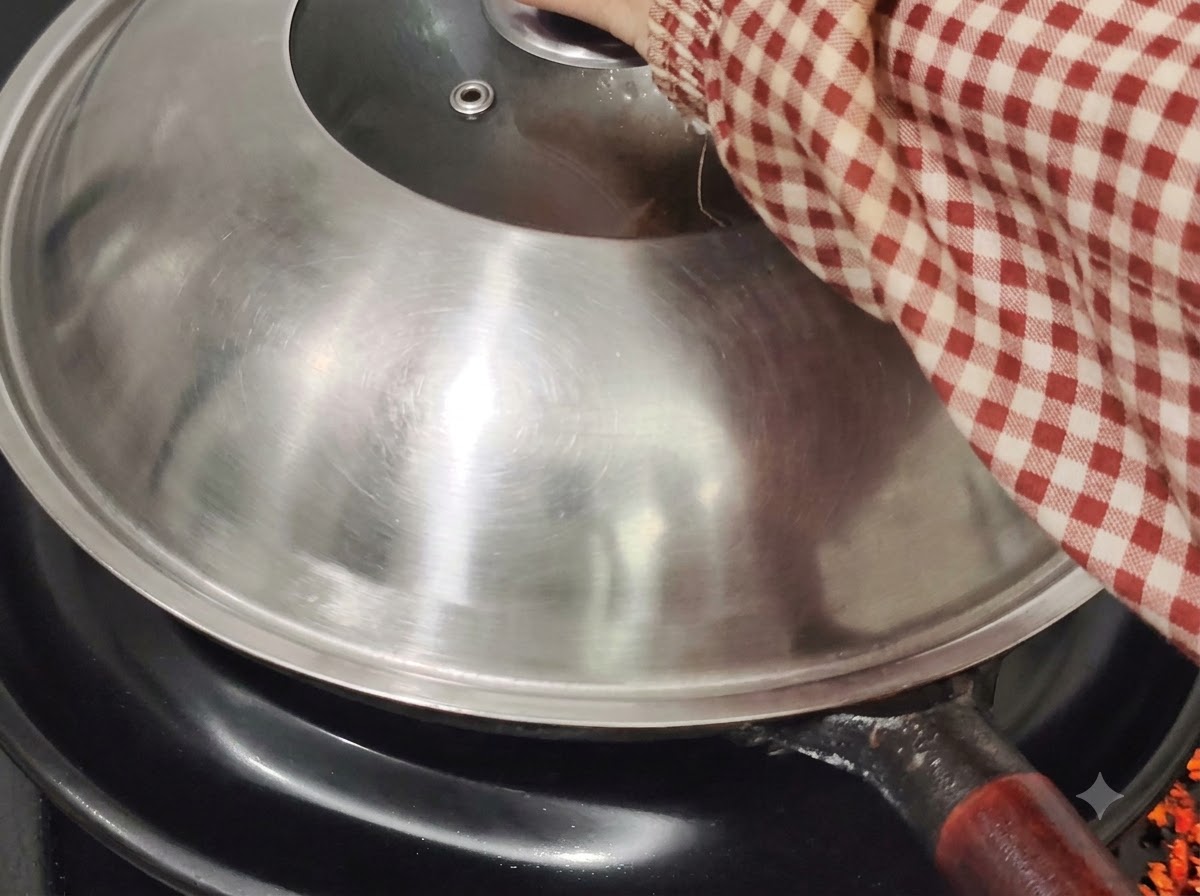 Couvercle de wok en métal posé sur le wok sur une plaque de cuisson, tenu par une main portant une manche à carreaux rouges et blancs.