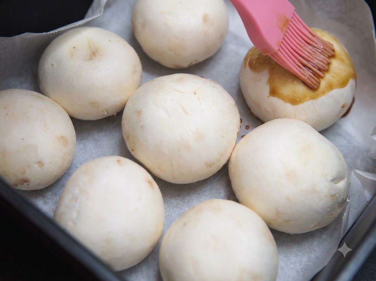 Un pinceau à pâtisserie en silicone rose appliquant de la sauce soja au miel foncée sur des chapeaux de champignons de Paris placés sur du papier sulfurisé à lintérieur dun panier de friteuse sans huile.