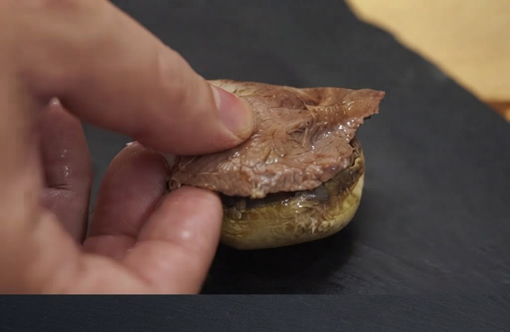 Une main plaçant un morceau de viande cuite sur une base de chapeau de champignon cuit.