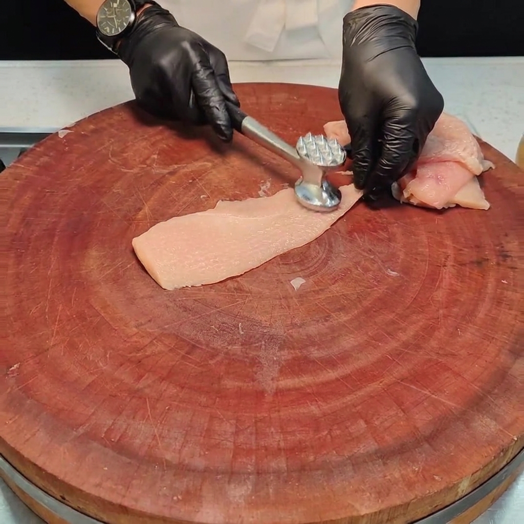 Un morceau de poitrine de poulet battu à plat avec un maillet à viande en métal sur une planche en bois.