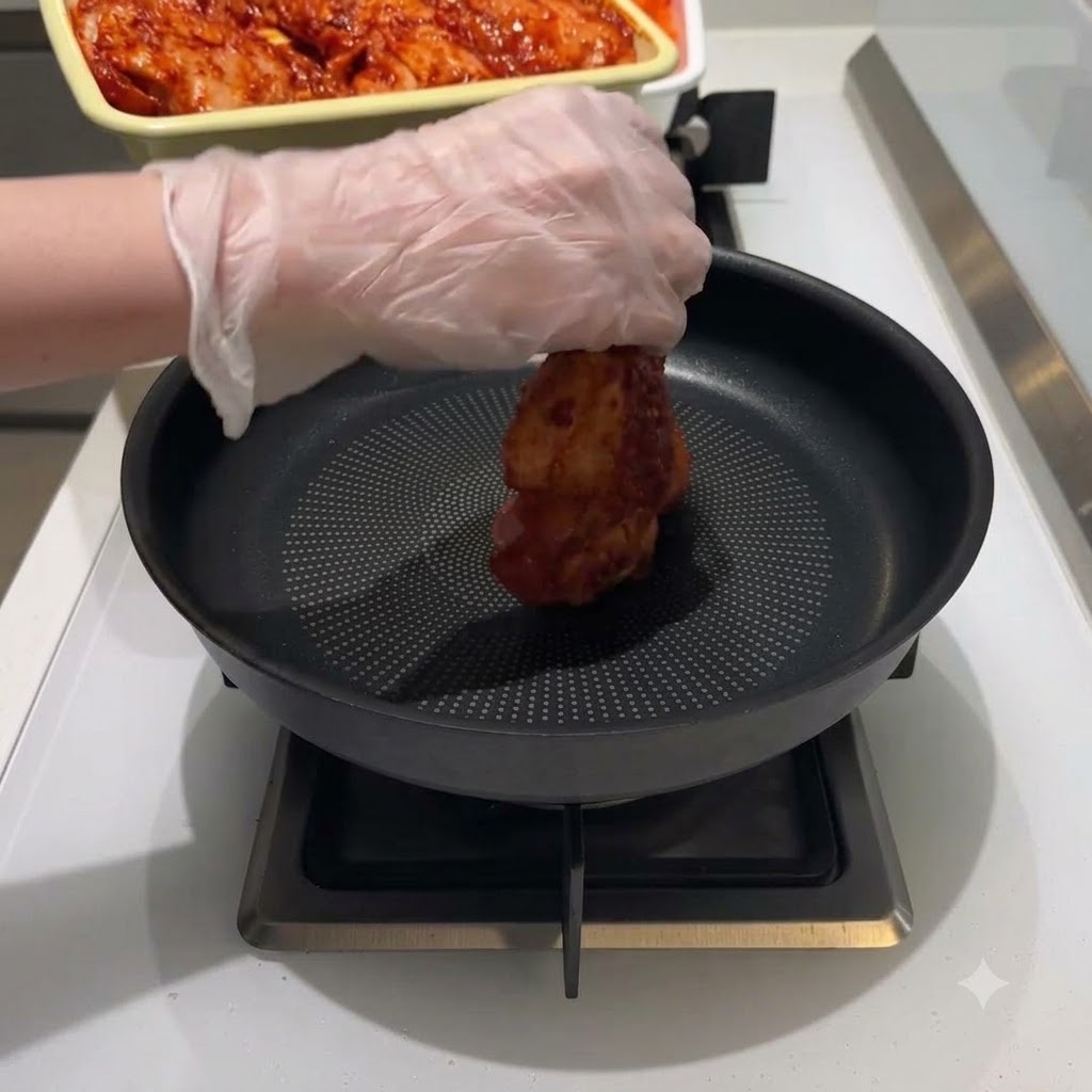 Une main gantée plaçant un morceau de poulet mariné rouge au milieu dune poêle à frire noire.