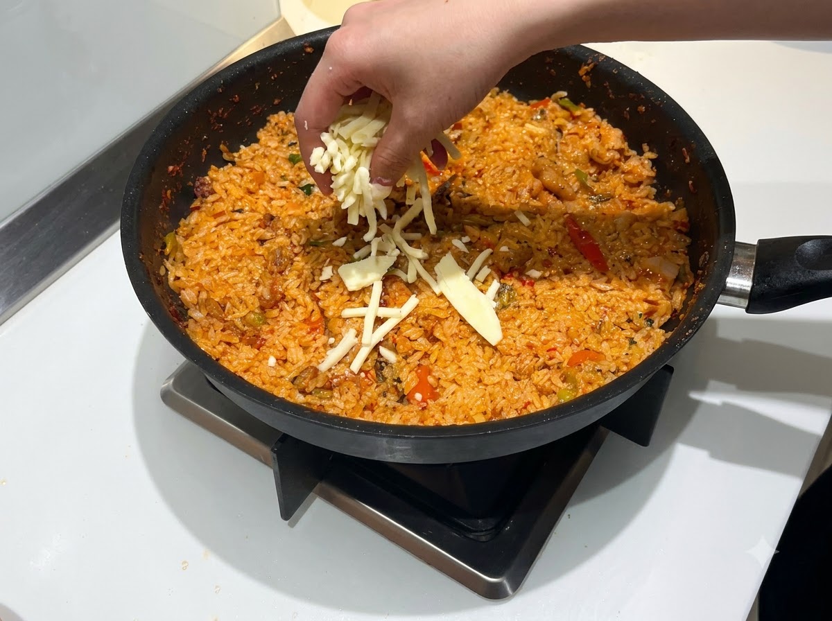 Une main saupoudrant une quantité généreuse de fromage mozzarella râpé sur une poêle de riz sauté rouge et épicé.