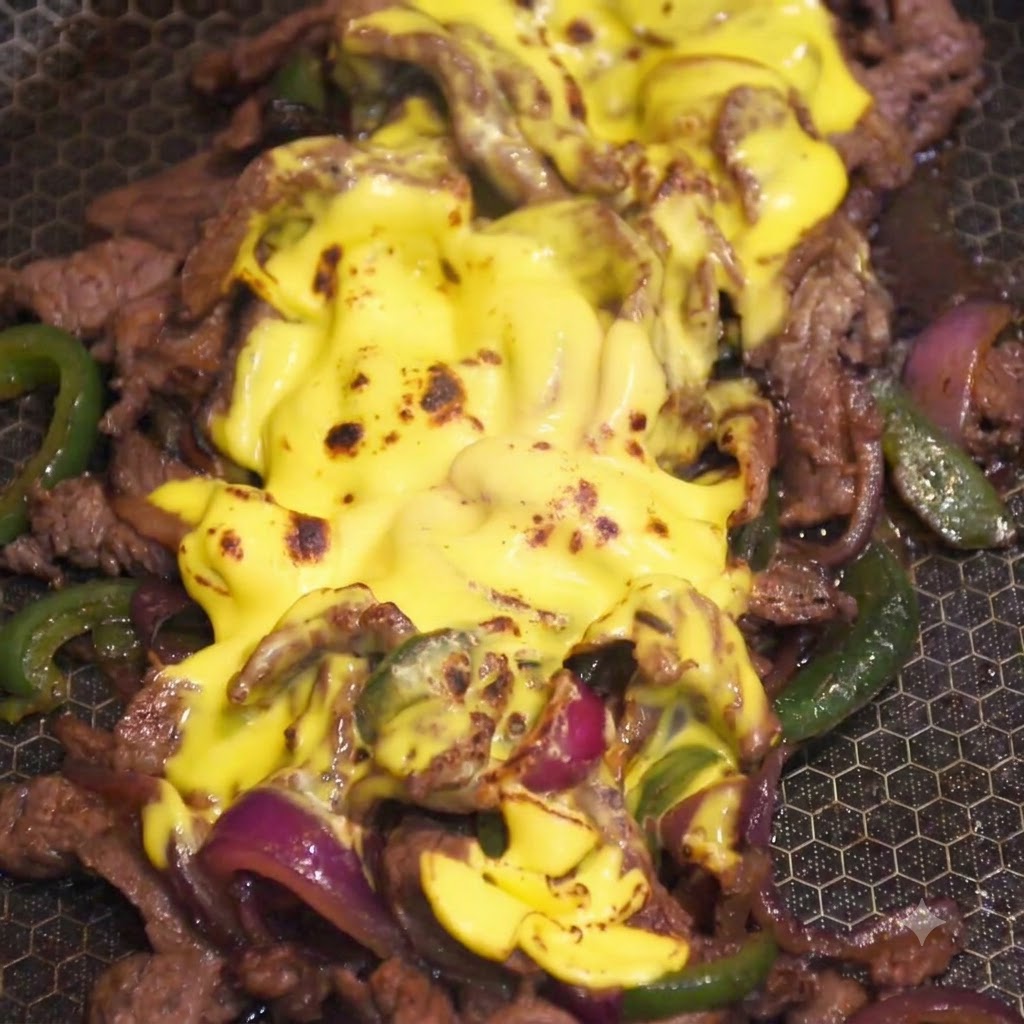 Un chalumeau de cuisine torchant la couche de fromage jaune fondu sur le mélange de bœuf et de légumes dans une poêle.