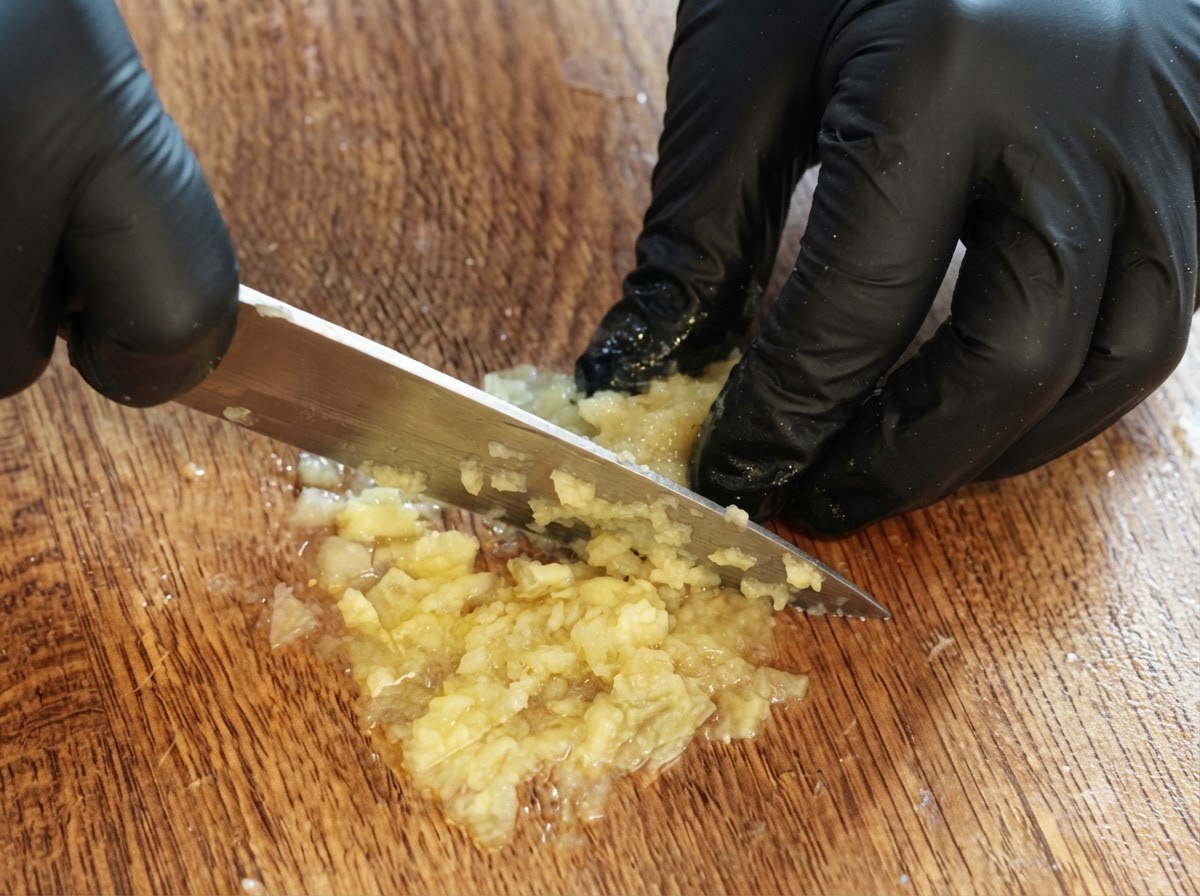 Une paire de mains gantées utilisant un couteau pour couper et réduire en purée les restes de chair de citron sur une planche à découper en bois.