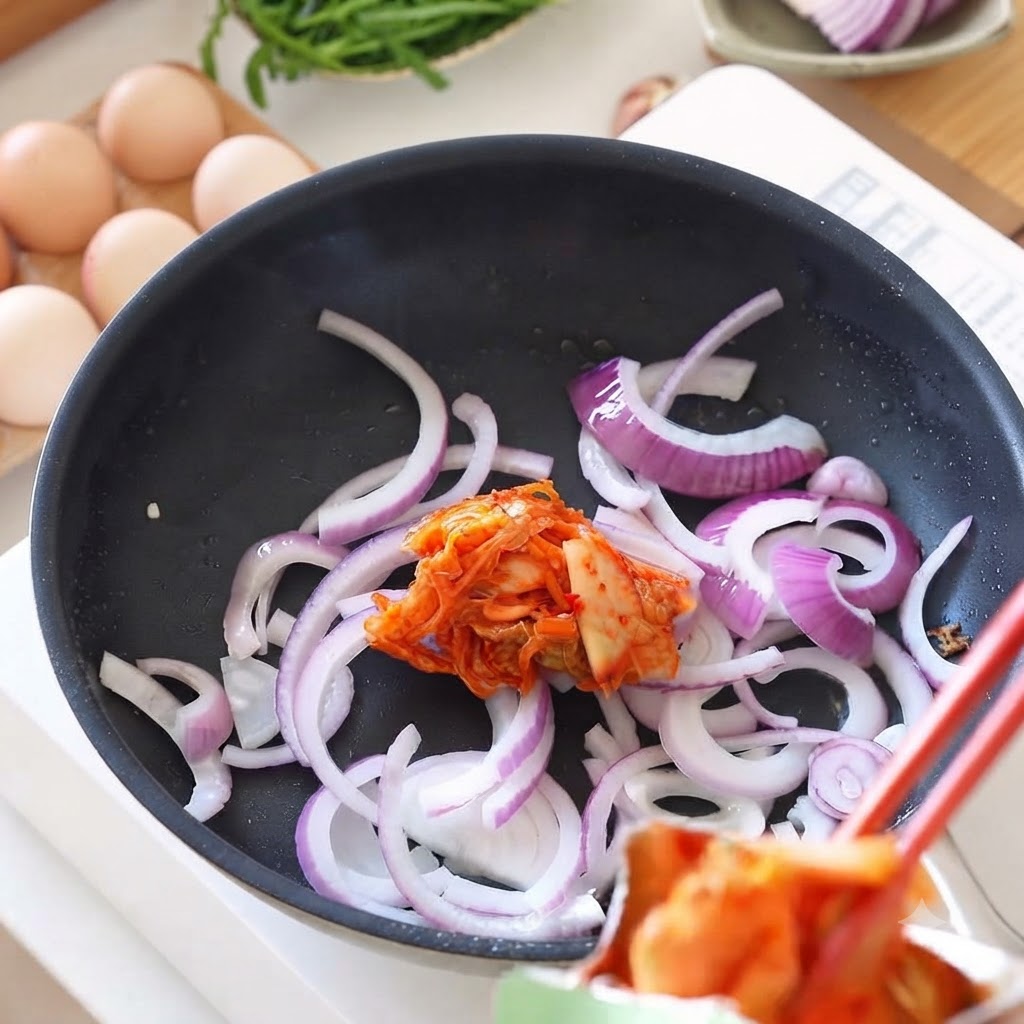 Du kimchi frais ajouté avec des baguettes dans une poêle contenant des oignons sautés.
