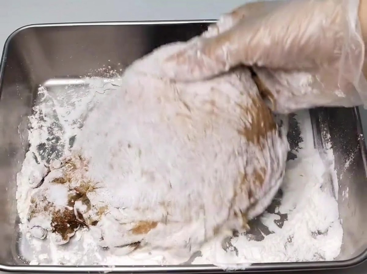 Une main gantée enrobant une cuisse de poulet marinée dune couche de fécule blanche à lintérieur dun plateau en métal.