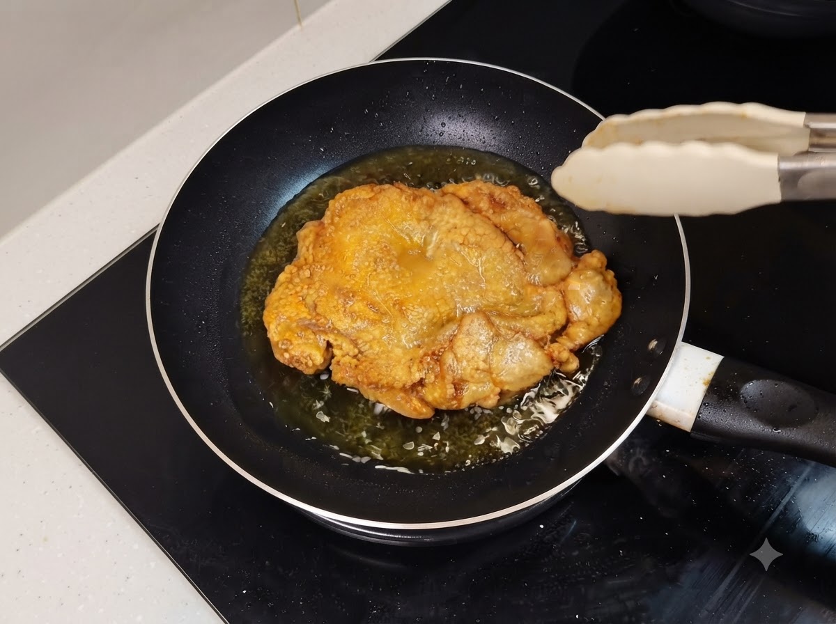 Une cuisse de poulet frite et dorée grésillant dans une poêle huileuse avec des pinces de cuisine à proximité.