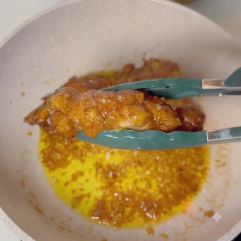 Des pinces tenant un pilon de poulet glacé au-dessus dune poêle de sauce au beurre à lail frémissante.