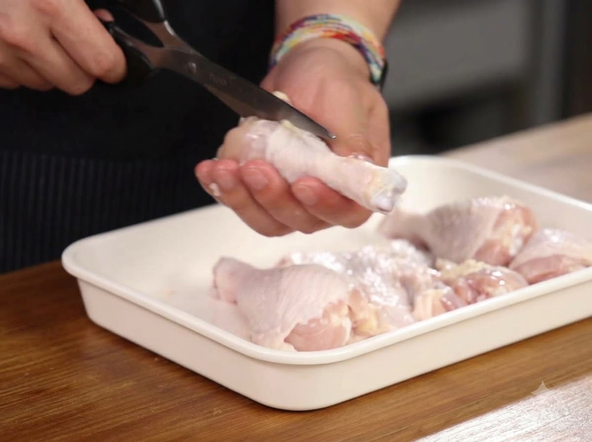 Mains utilisant des ciseaux de cuisine pour couper autour de los dune cuisse de poulet à lintérieur dun plateau blanc.